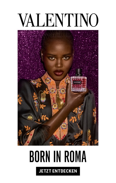 Ein Parfümflakon „Born in Roma“ von Valentino wird von einer schwarzen Frau in einem kunstvoll bestickten Outfit vor einem glitzernden violetten Hintergrund gehalten.